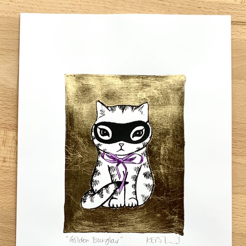 A screenprint: A masked kitten with a purple bow framed in a gold blocked square