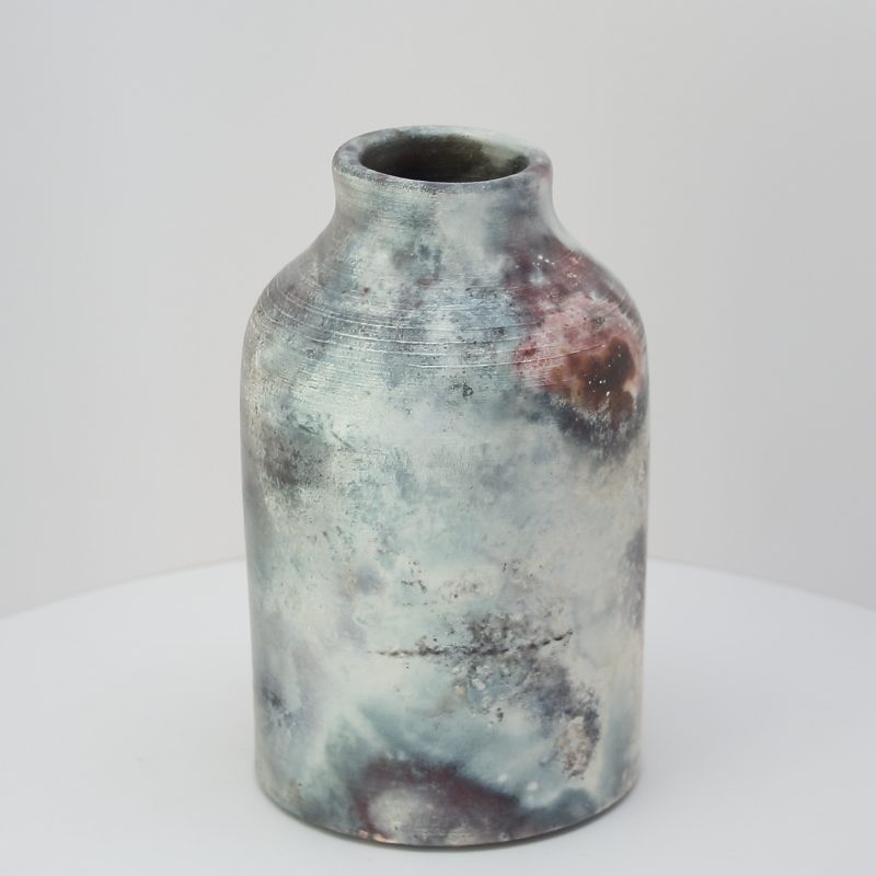 This wide mouthed bottle was wood fired for a day in an open pit after a copper solution and some copper wire had been applied.  The resulting marks on the pot are from the wood smoke (blacks and greys) and the copper (reds and black lines).