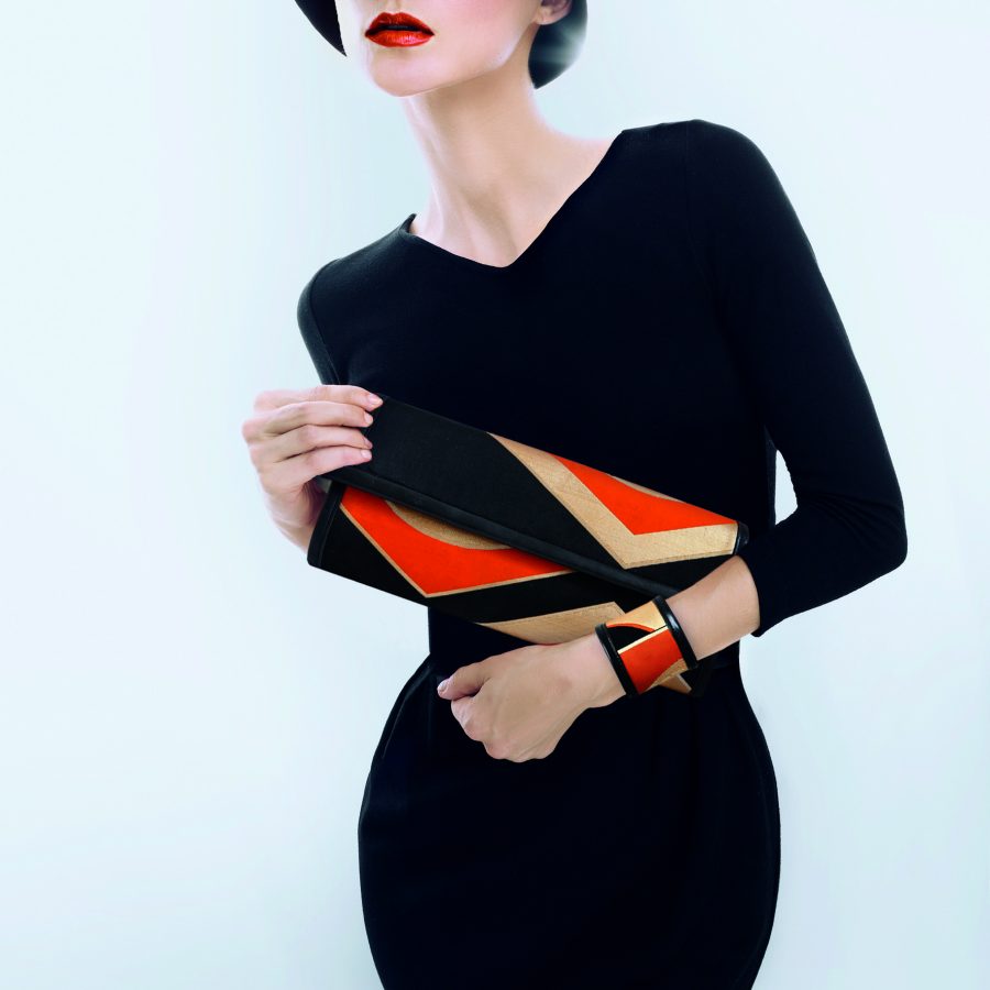 Elegant model wearing simple black dress, impacting orange lipstick, showcasing a geometric black gold and orange clutch bag and matching cuff bracelet