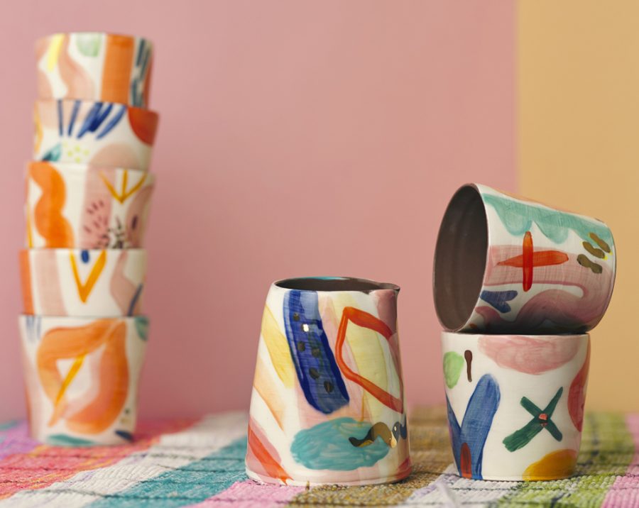 6 handpainted colourfully painted drinking vessels and one jug, all made in terracotta. the vessels are set on a stripy pink and green blanket and a backdrop of bubblegum pink card.