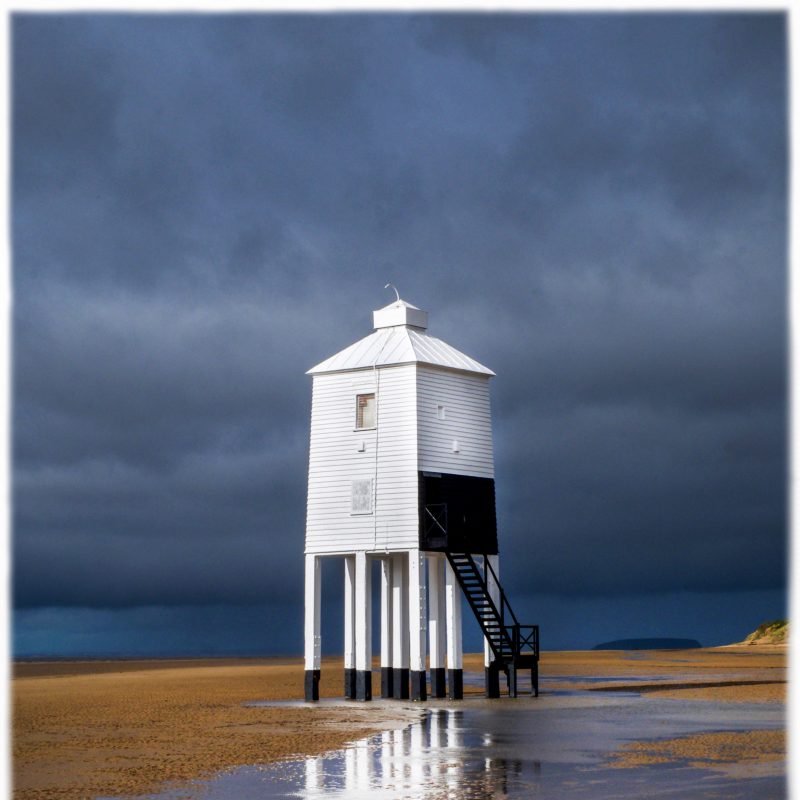A white wooden lighthouse on stilts on a sandy beach with stormy skies and the sea behind