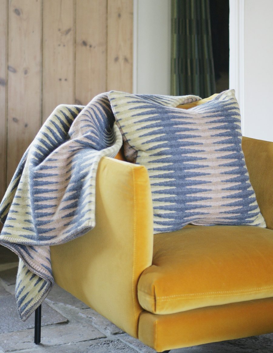 A woven throw and cushion in zigzag pattern draped on yellow velvet chair. The colours are greys and ochres.