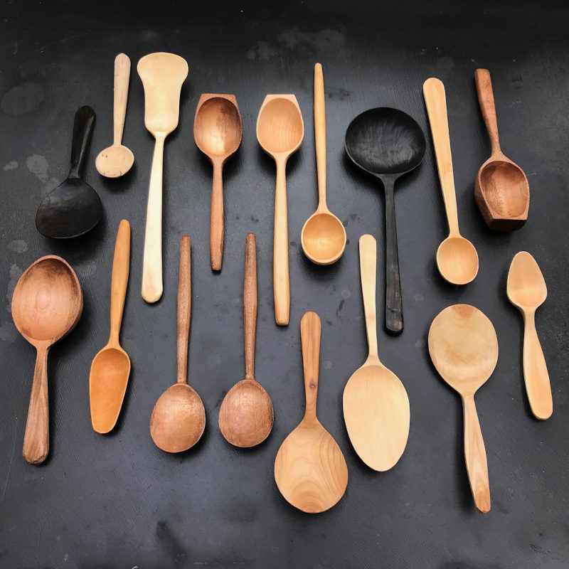 on a black background sit 17 hand carved spoons in various shades of natural wood.