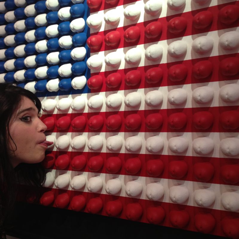 An American flag collage made from hundreds of casts of penis tips, with a woman licking one of the casts 