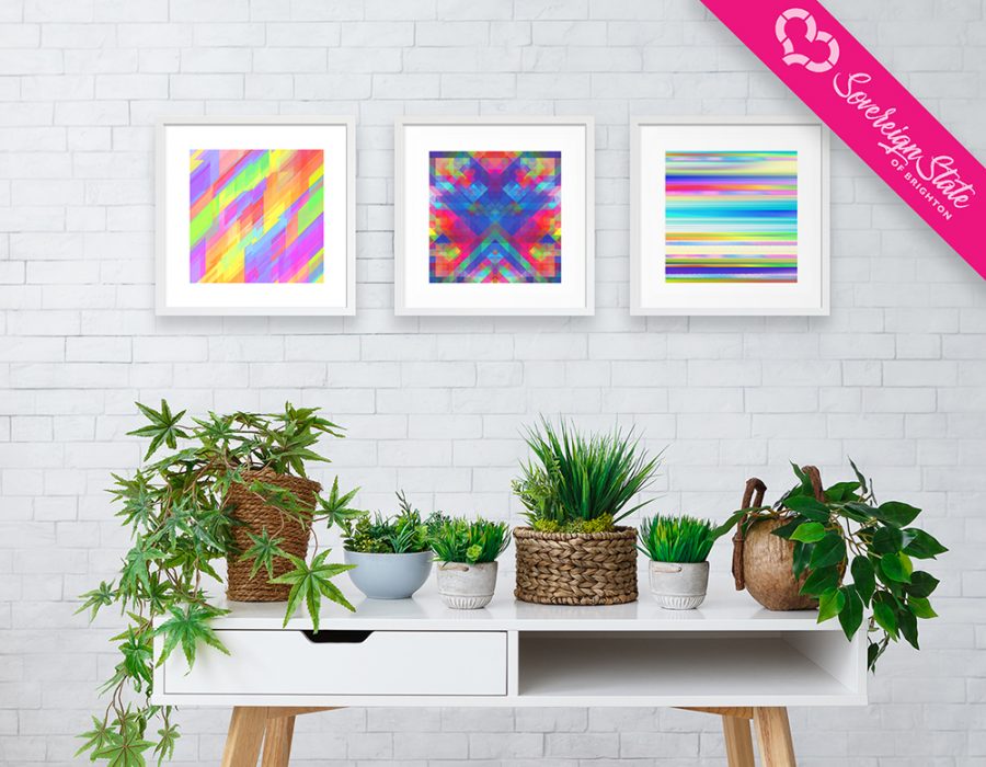 Three geometric square artworks in white frames hung onto a white brick wall situated above a white table with a variety of house plants on top