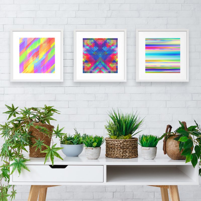 Three geometric square artworks in white frames hung onto a white brick wall situated above a white table with a variety of house plants on top