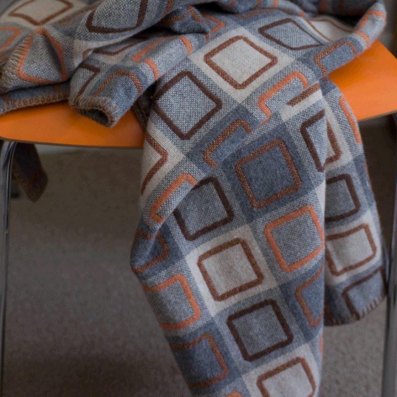 A plaid throw with orange and brown squircle motifs on soft greys and creams. Here shown on orange chair.