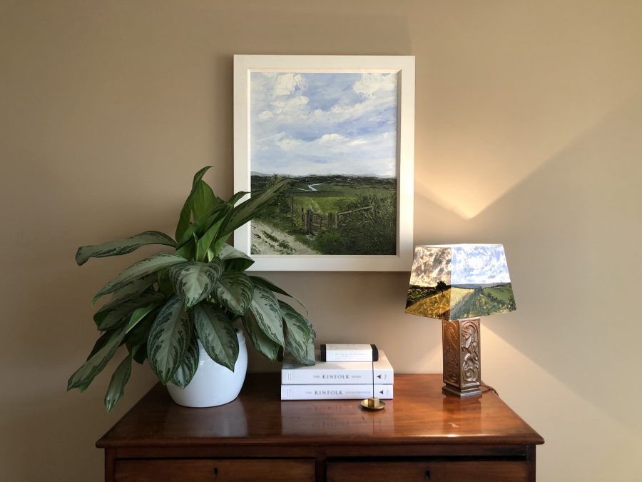 An in situ photo of a clay coloured wall with a centrally positioned painting in a white frame. Beside it there is a plant, a stack of books and an antique brown ceramic lamp with a hand painted rectangular lampshade. The lampshade features panoramic views of locations from around Roedean, Devil's Dyke and Southwick.  The textured palette knife landscape painting pictures Amberley Mount. Soft clouds and a light blue sky contrast with spring greens of the rolling hills and a waving river. In the foreground, the view is framed by a wooden gate and the South Downs Way path in the bottom left corner.