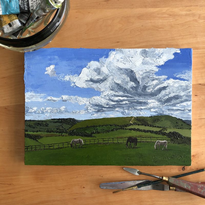 Painting of three horses grazing on a sunny September day near The Jack and Jill Windmills. Rolling hills of the South Downs feature in the background with a large, dramatic cloud above. 