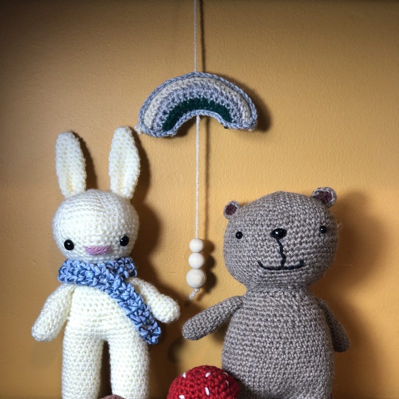 A selection of crocheted stuffed animals; a brown bear and a white rabbit with a blue scarf, a red toadstool and brown porcini mushroom, a rainbow hanging with grey, cream and green stripes, with 3 wooden beads hanging down from it. A products range from 6cm to 26cm tall and 5cm to 15cm wide