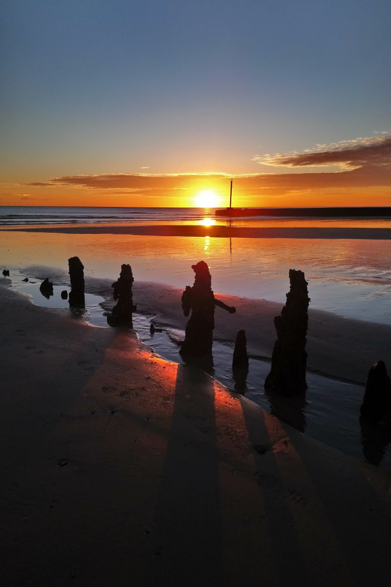 Photograph of a sandy beach and wooden groynes at sunset with a low tide.  The sun comes between two groynes highlighting the shallow water at their Base. 