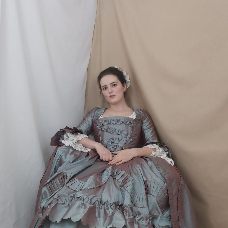 Model is seated and reclining wearing a voluminous eighteenth century gown made of grey green taffeta shot with red, it is heavily trimmed with ruffles and lace. Her hands are resting in her lap and looking directly into the camera, her hair is up and she is wearing a white cap on the back of her head.