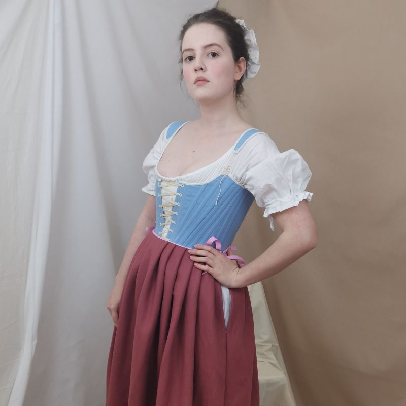 Model stands facing three quarters towards the camera with a hand on her hip and looking down at the camera slightly. She is wearing eighteenth-century clothing including a pair of blue lace up stays (similar to a corset), a dark pink wool petticoat, with a white shift with short puffed sleeves underneath. Her hair is up and she has a white cap on the back of her head.