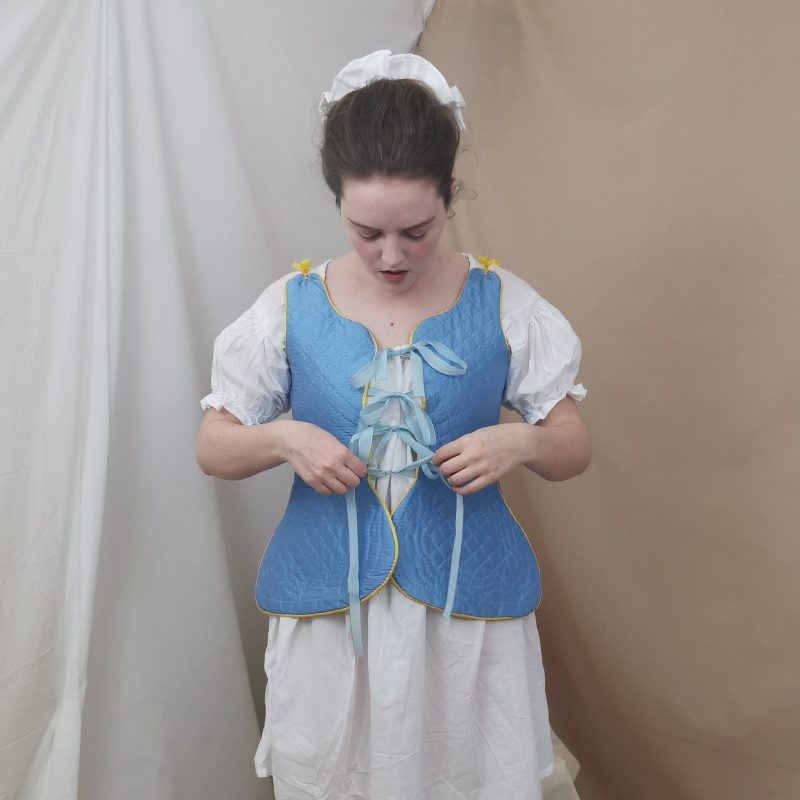 Model is standing looking down at her hands which are tying a bow in the front of her waistcoat. She is wearing eighteenth-century undergarments consisting of a light blue silk quilted waistcoat with ties down the front to secure it shut, underneath it she wears a white linen shift with short puffed sleeves. Her hair is up and she has a white cap on the back of her head.