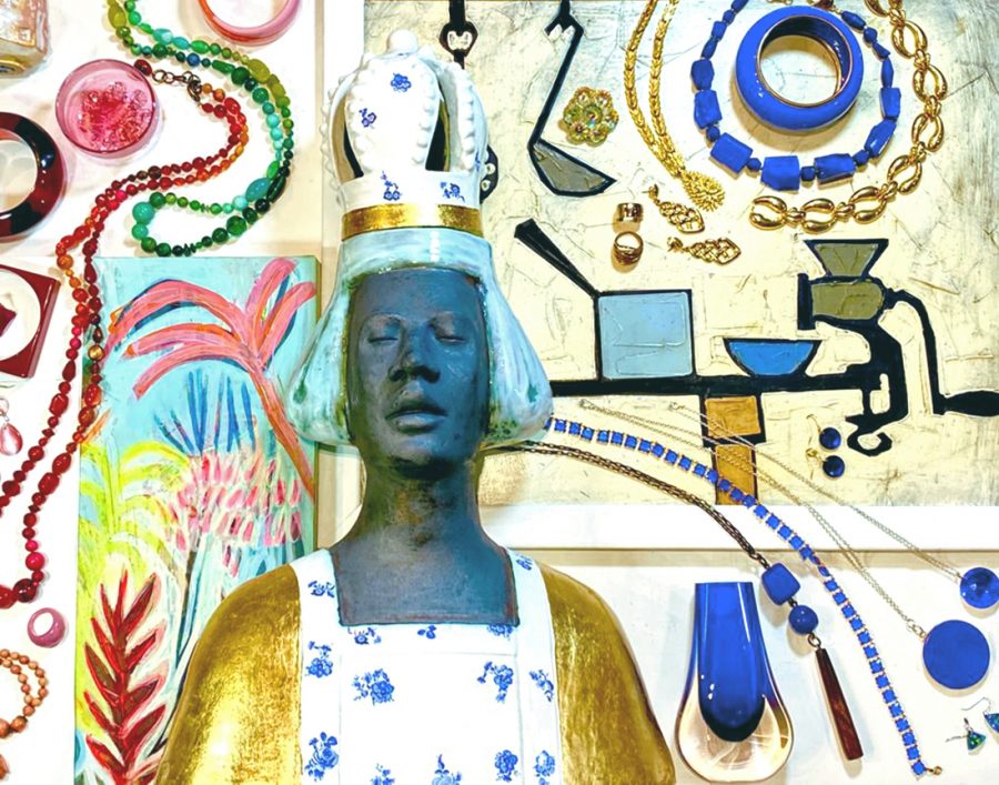 A photograph taken from above of a ceramic sculpture head with gold leaf and blue flowers transfers. laying on top of two different original paintings. One is brightly coloured palm images and the other shows naive style kitchen equipment. All of the artworks have coloured and golden jewellery laid on top and around plus some coloured glass pieces.  