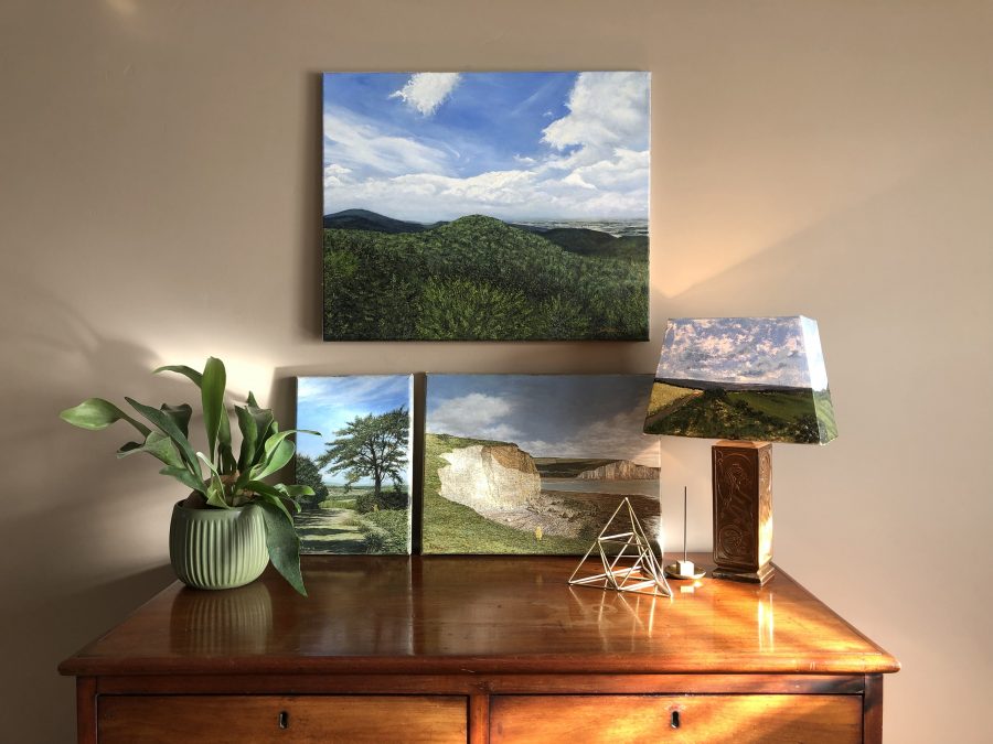 A cluster of landscape paintings featured against a clay colour wall, above a dark wooden chest of drawers. Top painting features a view from the top of Kalenica Mountain in Poland that spans wooded hilltops. Underneath it, there are two paintings. The smaller one is of a yellow teddy waving at walkers in Ashdown forest. The teddy named Will is standing under a tree on the side of a path. In the other painting, Will is standing at Hope Gap on the Sussex Coast at Seaford, overlooking cloudy sky and chalky cliffs at low tide. To the right, a lamp with painted lampshade of a landscape of sea view overlooking Roedean. 