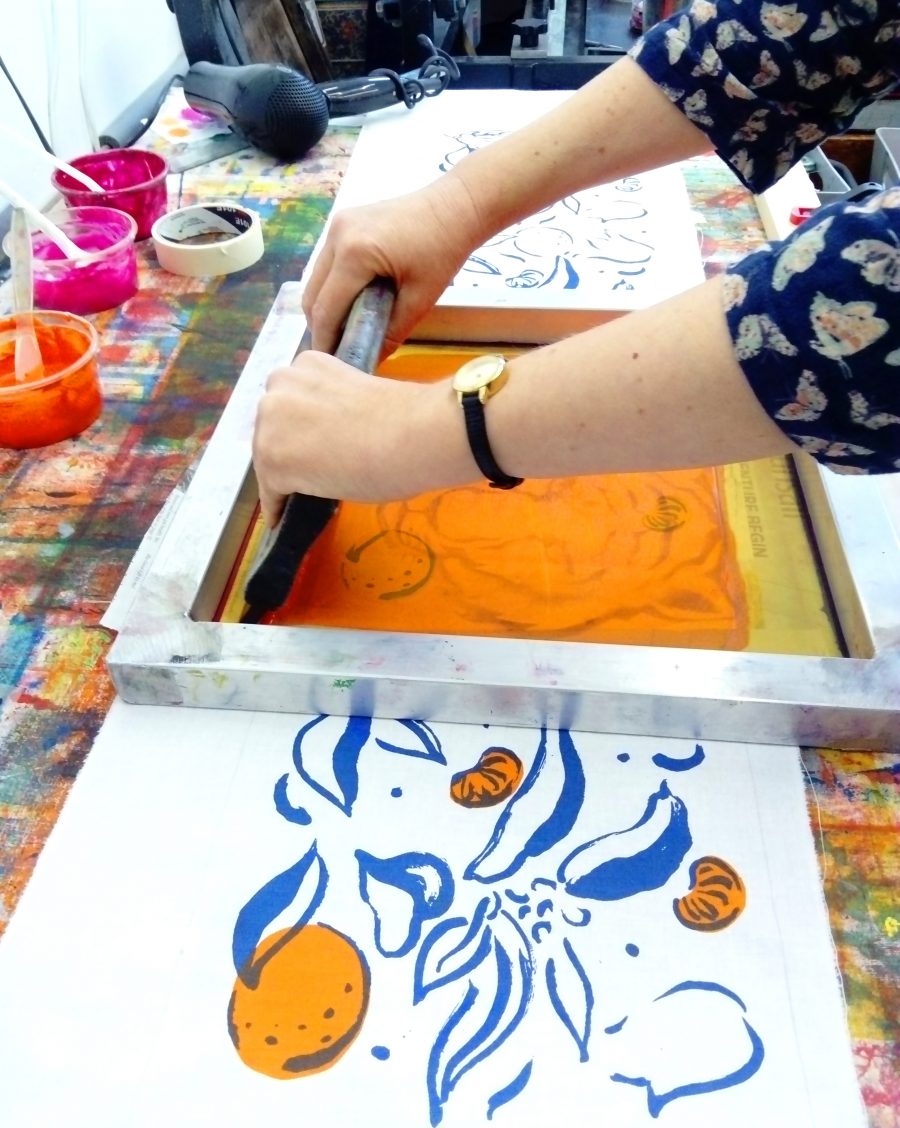 screenprinting in action shot showing hands holding a squeegee to screen print a blue and orange hand drawn image of leaves and fruit onto a length of fabric.