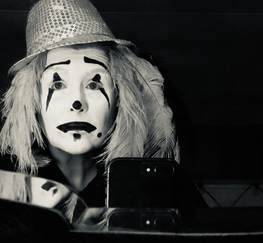 black and white self-portrait as white-faced clown, black emotion lines & clown nose, feathers for hair, a spangly jaunty hat; sorrow.