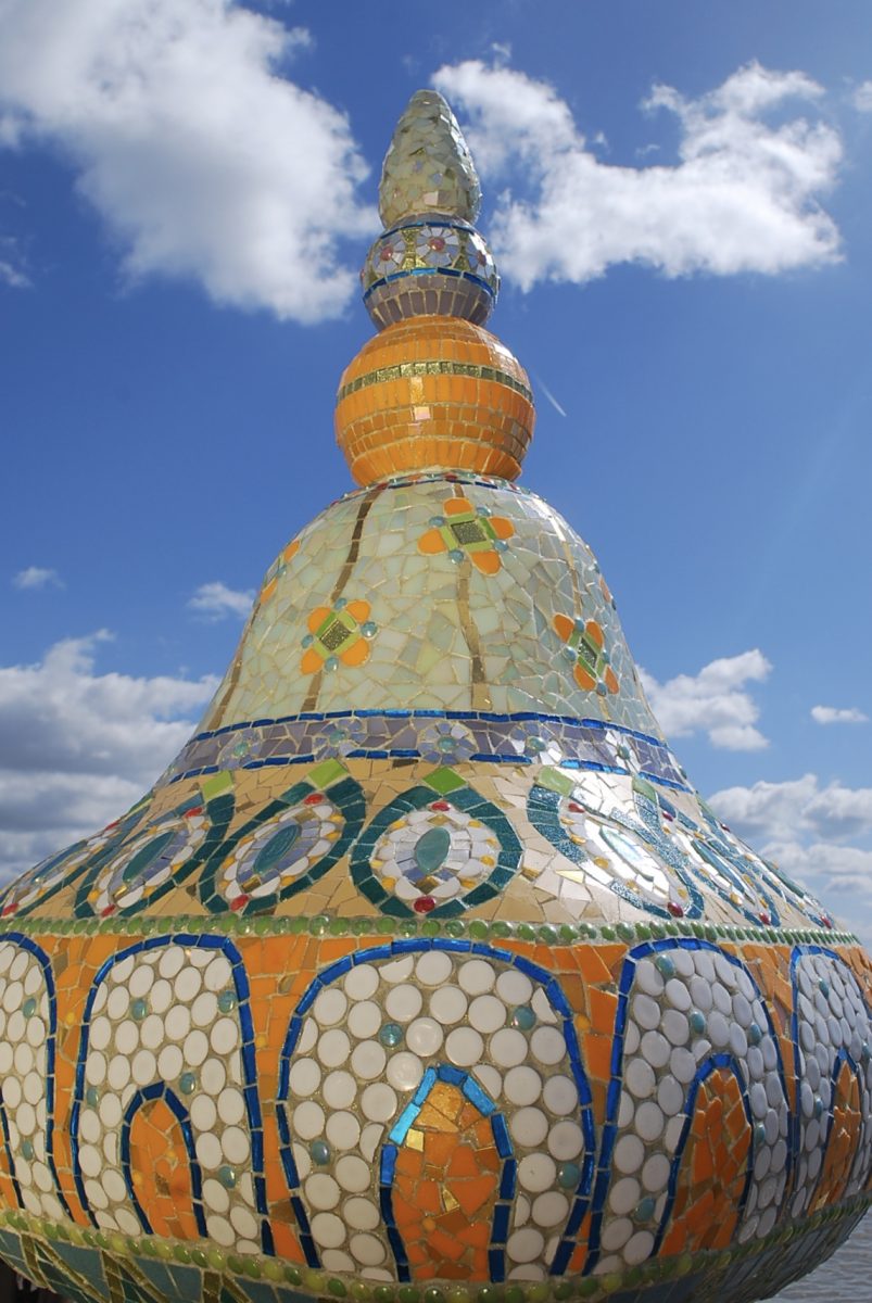 An architectural shape with a circular top and pinnacle. Clad in glass, hand cut mosaic tiles. Greens, turquoise, orange and golds. 