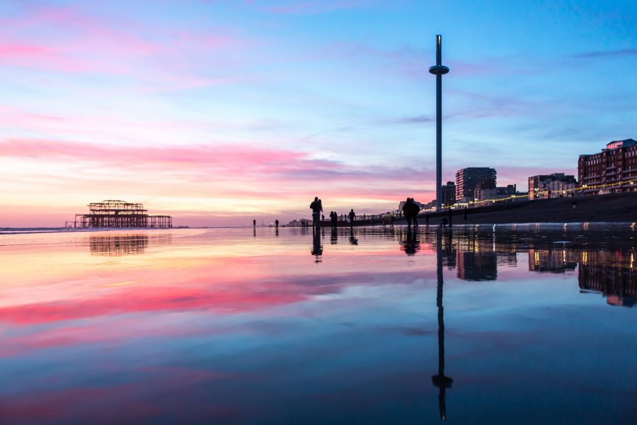 A photograph of the BAi360 slowly ascending to the skies, with a sunset view across the beach from the Palace Pier, clear hues of pink and crimson cloud, walkers on the beach in silhouette.
