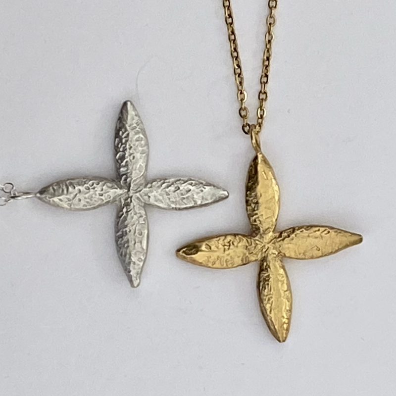 A close up photo of a silver and a gold cross shaped pendant on chains. The pendants are a hammered texture and the 4 prongs of the cross are shaped like petals