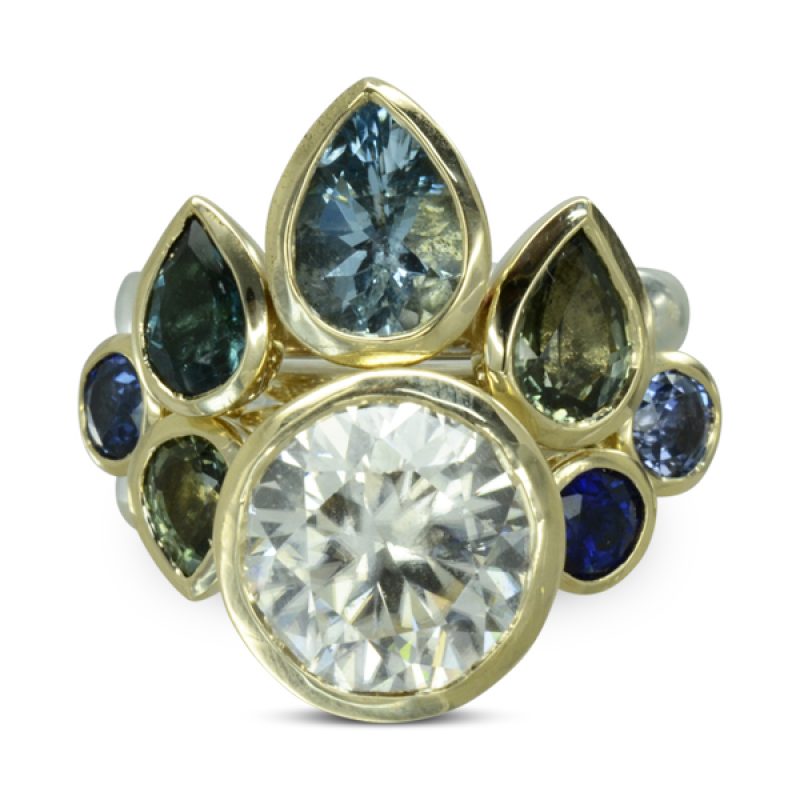 A Sapphire Peacock Stacking Cocktail Ring in platinum with 18ct yellow gold settings. A three carat white sapphire mixed with teal, indigo and green sapphire pear shapes and rounds. The ring comes apart into three, the outer rings can be worn alone and when they are together are locked into place by the outer sapphires on the middle band.