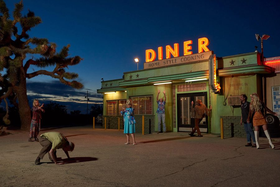 An external of an American diner at nighttime in the desert, with a foreground of characters behaving strangely. 