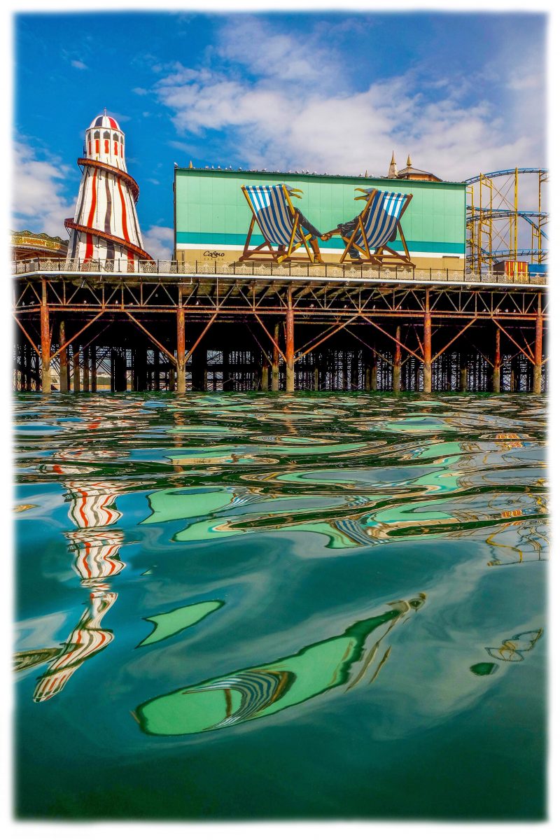 The heater skelter on Brighton pier with a large painting of two seagulls sitting on deck chairs below the pier is the reflection of the colours of the pier, the sea is very calm. 