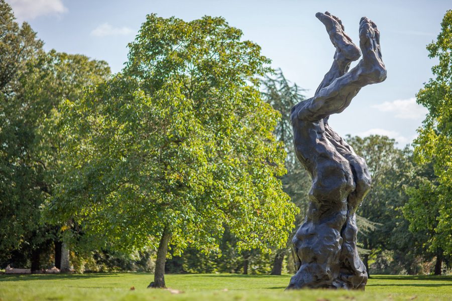 Sculpture of legs emerging from the ground from the waist downwards, the feet being at the top of the sculpture. The legs are the same height as the tree the sculpture is placed next to.