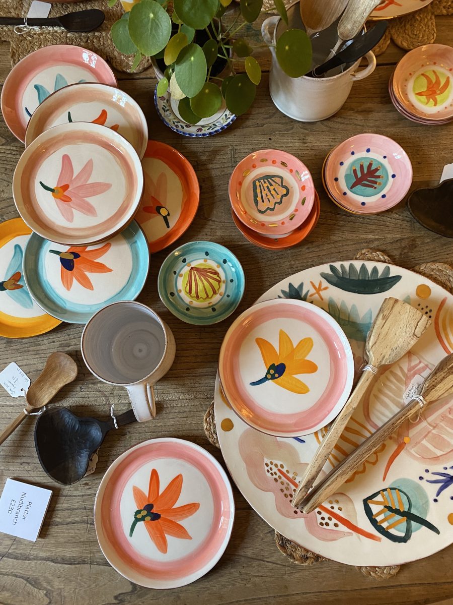 top down view of pine table, you can see brightly coloured hand painted plates and platters as well as hand carved spoons, at the top is a money plant.