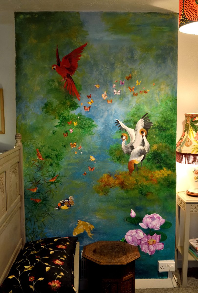 This mural depicts a dreamy tropical river and cloud forest where birds and butterflies fly above fish and lotus flowers.