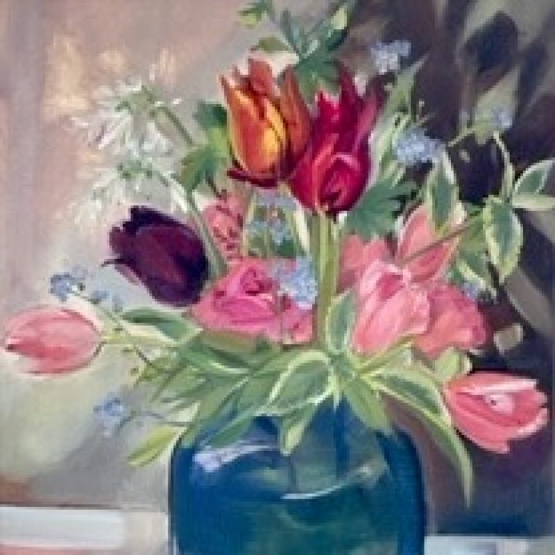 Blue vase with a pink, purple and orange tulips. Variegated leaves with small blue and white flowers enhancing the tulips. Shadows cast on the wall behind in blues and purples. Pink and blue light shadows on the tablecloth