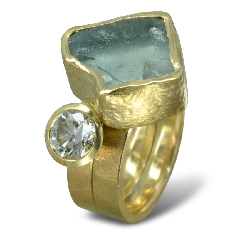 Aquamarine Chunk Diamond Stacking Dress Ring handmade in recycled yellow gold with a half carat recycled round brilliant diamond and an 18 mm AAA rough aquamarine gemstone. The metalwork has a rough hammered texture and the bands are 3mm in width. The Diamond and Aquamarine sit on top of the bands and are in bezel settings where the gold goes all the way around the stones..
