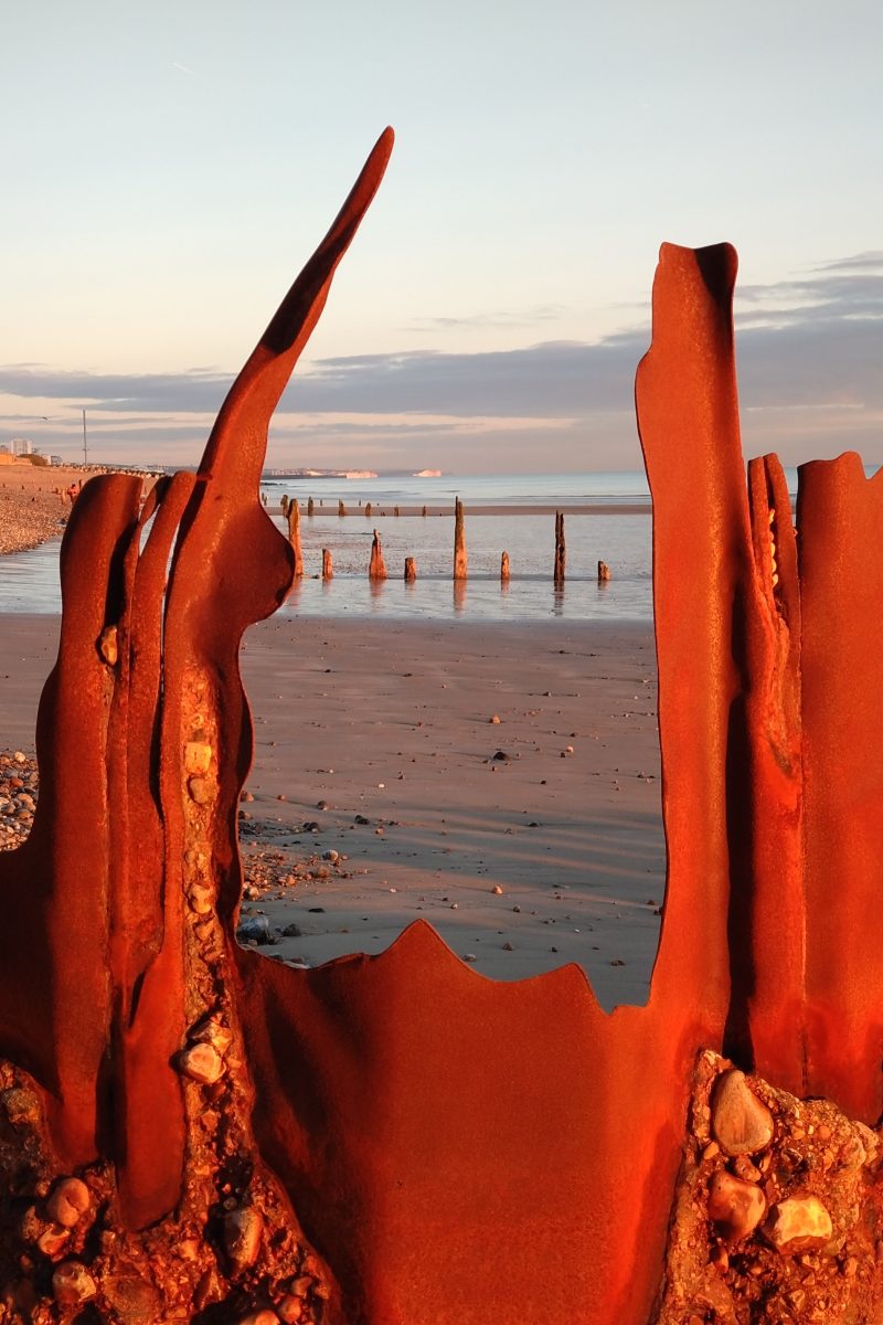 Photograph of a metal groyne, sculpted by the sea, rusty and glowing at sunset, through which a sandy beach and wooden groynes can be seen