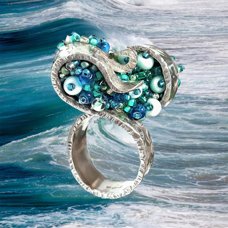 Swirled silver statement ring with aqua and blue beading on an image of sea waves.