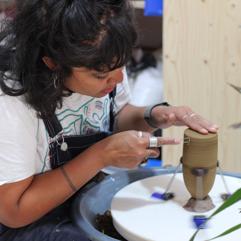 Female ceramicist at the potters wheel, working on an up-side-down bottle. The face is close to the work, the hands with lots of silver jewellery holding tools to shape the sand coloured clay.