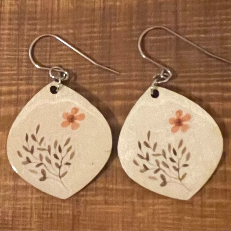   Wooden drop shape earrings with a small orange flower and leaves on each of then. Resting on a wooden board 