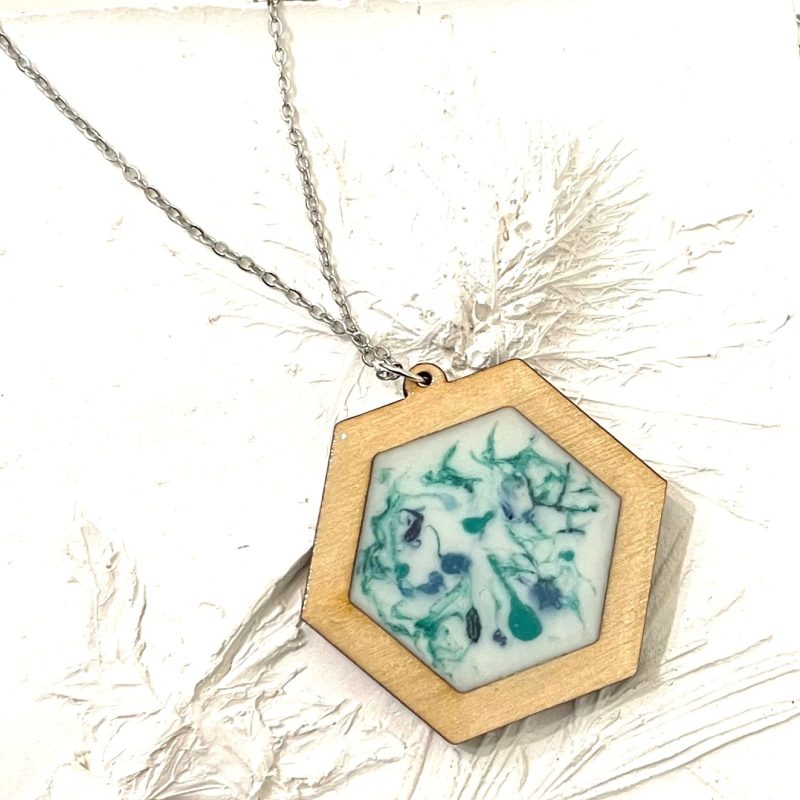Wooden hexagon shape pendant with blue, green and white resin in the centre attached to a stainless steel chain. On a white background made from plaster of Paris that has impressions of a thistle in it. 