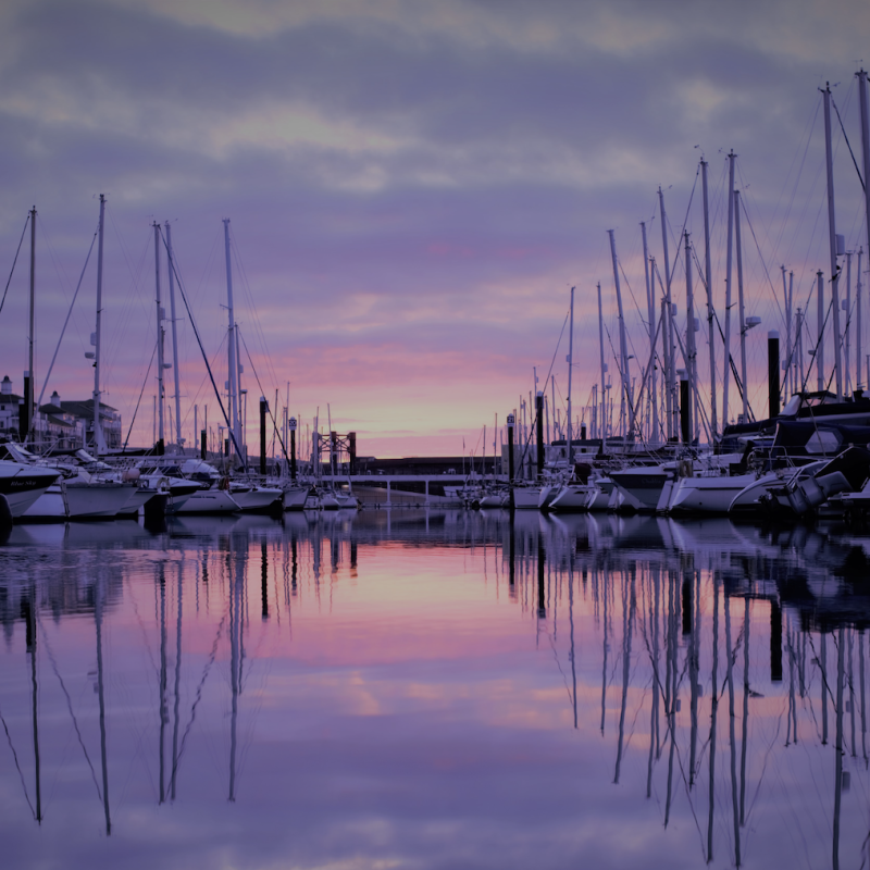 A sunrise image of boats on the water at Brighton Marina. Colours of pinks and purple of the sky reflect in the water 