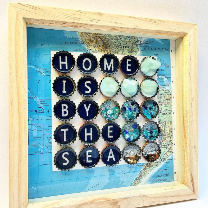 Wooden Box frame with recycled atlas map background, recycled bottle tops mounted on to the background filled with recycled materials (blue sequins, recycled nature magazines & miniature pebbles) with the words ‘Home is by the Sea’. 