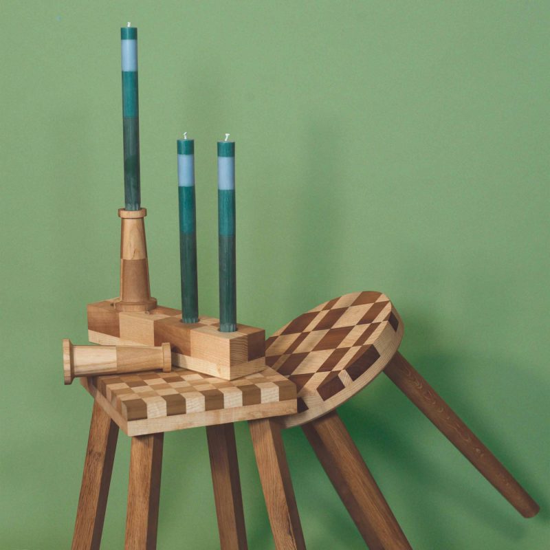 Two wooden checkerboard stools, two single candlestick holders and one block candlestick holder with green candles set against a green background
