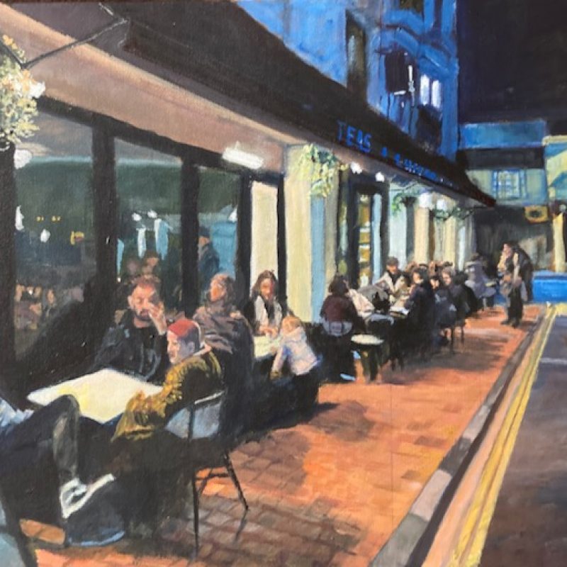 A view of the outside of a cafe in the evening. Customers are sitting at tables on the pavement