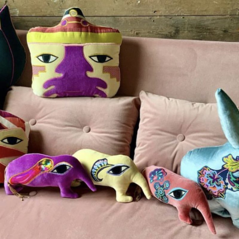 two brightly coloured Peruvian head cushion sit with 3 medical elephants and one rabbit. all these cushions are made out of vintage velvets and embroidery. sitting again pink velvet sofa.