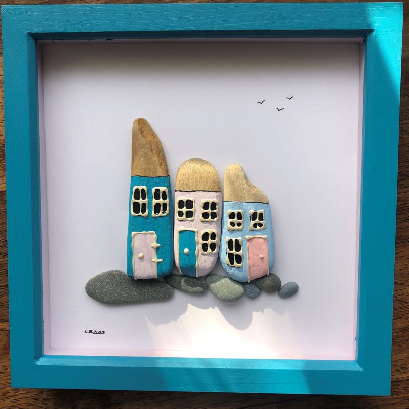 3 houses made of driftwood and painted bright blue, lilac and baby blue , sat on a row of smooth grey pebbles and framed in a 24cm x 24cm deep box frame that has been painted to match - bright blue with a lilac inner.