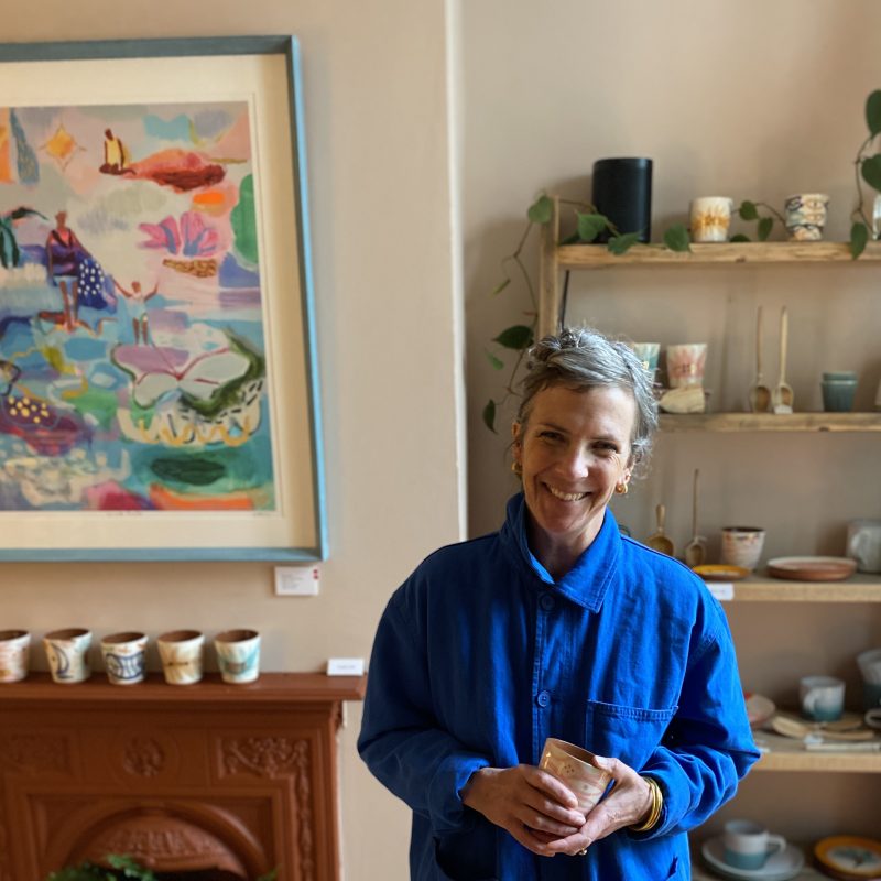 becky smiling,  dressed in blue and holding a piece of pottery. behind is a red fireplace with a print and pottery and a book case with more pottery and hand carved spoons.