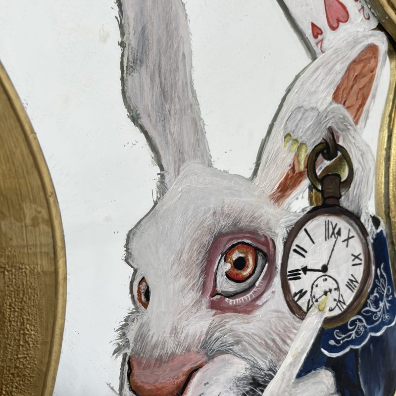 The White Rabbit from Alice In Wonderland is painted on to a small dressing table mirror. It is peering in from the side. The colours are rich with with red/orange eyes, in contrast with his blue suit. 