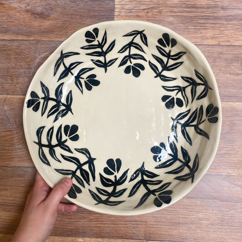 A birds eye view of a large ceramic platter on a wooden table top with hand painted black flowers around the edge and a hand holder the rim 