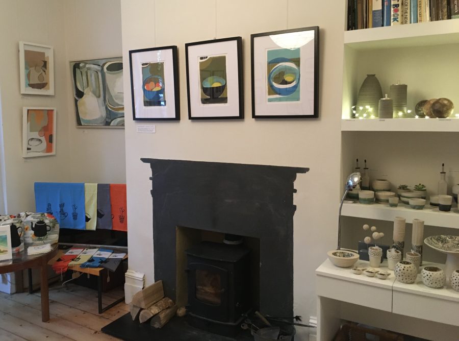 Living room interior with a dark fireplace surround in the centre and various framed artworks displayed on the wall and Ceramics on shelves in an alcove.
