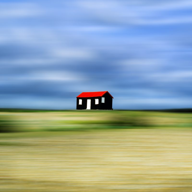 black hut with red roof on a pebble beach cloudy sky behind 