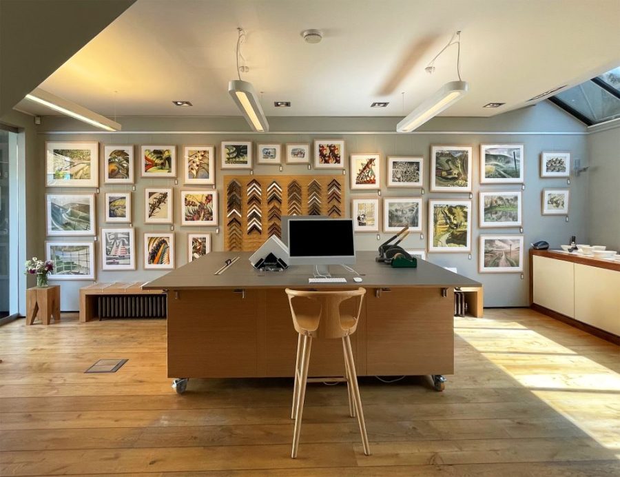 A gallery with framed prints on the wall. A table, chair and laptop is in the middle of the room. 